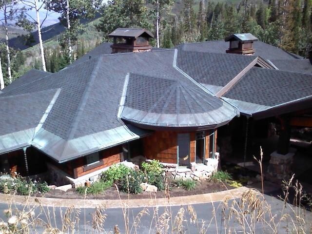 Large residential home with complex multi-peak roof and dormers, featuring wood siding and stone accents, surrounded by trees.